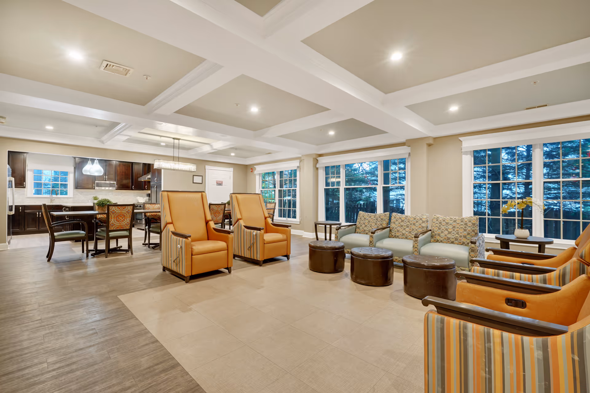 A spacious and well-lit common area in a senior living facility featuring comfortable seating including armchairs and sofas arranged around small round ottomans. Large windows allow natural light to fill the room, and in the background, there is a dining area with a table and chairs adjacent to a kitchen with dark cabinetry and modern lighting fixtures.