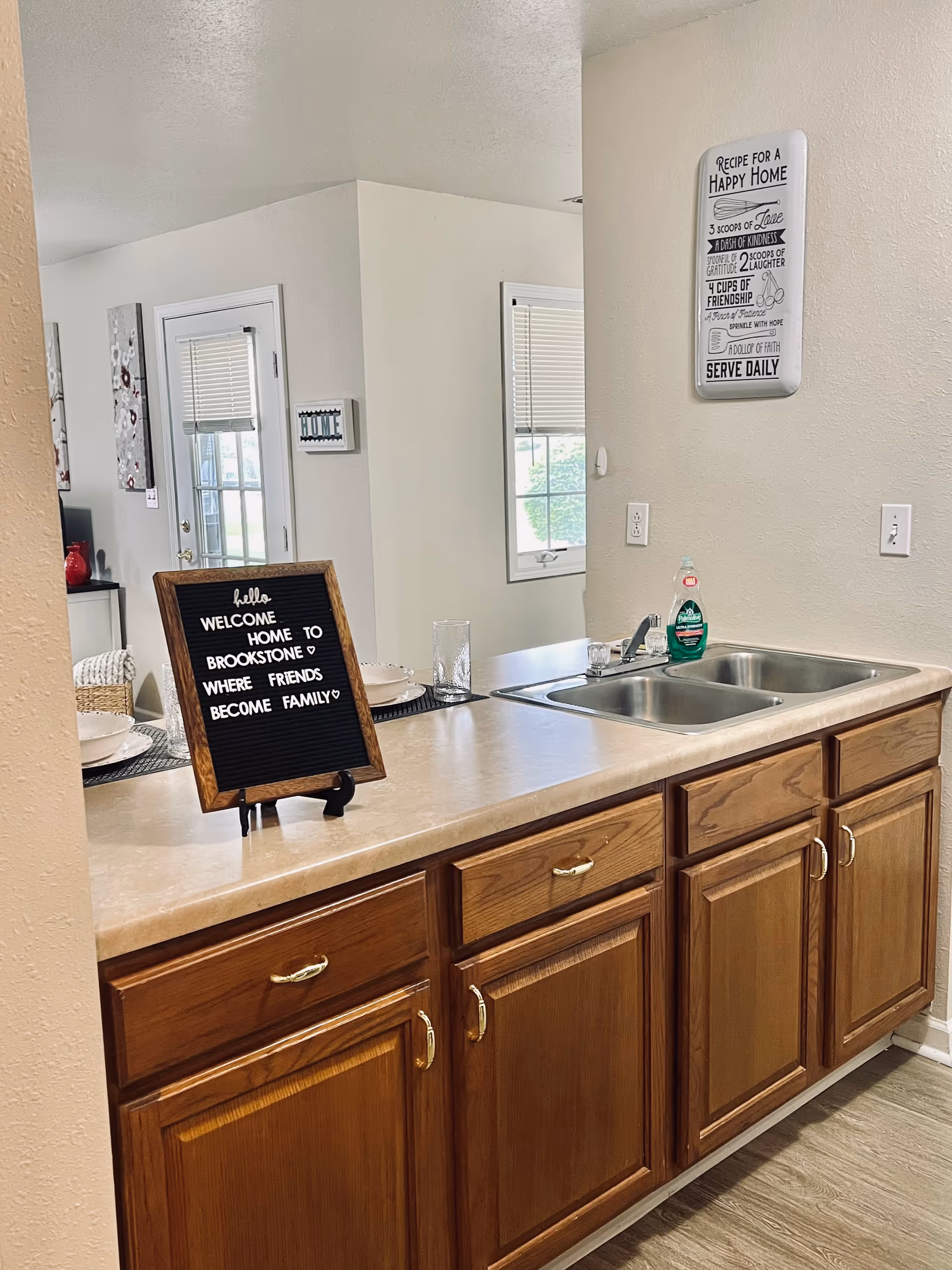 Interior view of a kitchen area with wooden cabinets and a double stainless steel sink. On the countertop, there is a small black letter board with a wooden frame that reads 'hello WELCOME HOME TO BROOKSTONE WHERE FRIENDS BECOME FAMILY'. A bottle of Palmolive dish soap and a glass are next to the sink. In the background, there is a window with blinds and a wall decoration with the text 'RECIPE FOR A HAPPY HOME' listing ingredients like love, kindness, gratitude, laughter, friendship, patience, hope, and faith. Part of a dining area with place settings and a door with a window is visible in the background.
