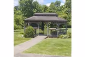 A wooden gazebo with a shingled roof situated in a grassy area with a concrete pathway leading to it. The gazebo is surrounded by green trees and bushes under a clear sky.