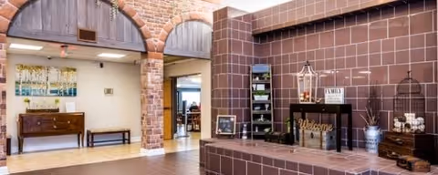 Interior view of a senior living facility lobby area with brick archways and walls. There is a wooden bench and a small table with a painting on the left side. On the right side, there is a tiled counter with decorative items including a lantern, a 'Welcome' sign, and a small shelf with plants. The space has warm lighting and a welcoming atmosphere.