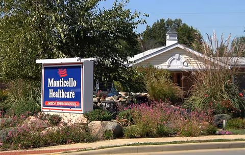 Monticello Healthcare exterior featuring a blue sign, landscaped garden, and a white building facade.