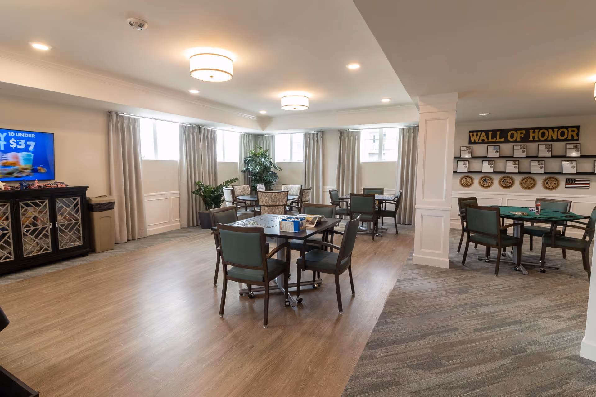 A well-lit common area with several tables and chairs arranged for group activities or dining. The room features large windows with curtains, indoor plants, and a 'Wall of Honor' display with plaques and decorative plates on the wall. There is a cabinet with a television on top showing a promotional image.