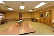 A spacious room with beige walls and carpeted floor featuring a large wooden table in the center with a small flower arrangement. There are several smaller tables and chairs along the walls, a window with blinds on the left, and double wooden doors on the right side of the room.