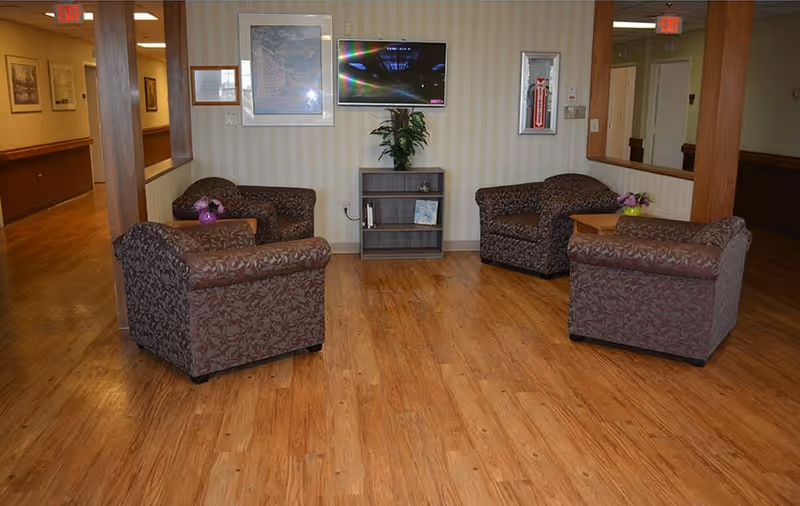 A seating area in a nursing facility with four patterned armchairs arranged around a small wooden table with a flower vase. A small bookshelf with a plant and books is against the wall beneath a mounted flat-screen TV. The floor is wooden, and the walls have light-colored wallpaper. There are hallways visible on either side of the seating area.