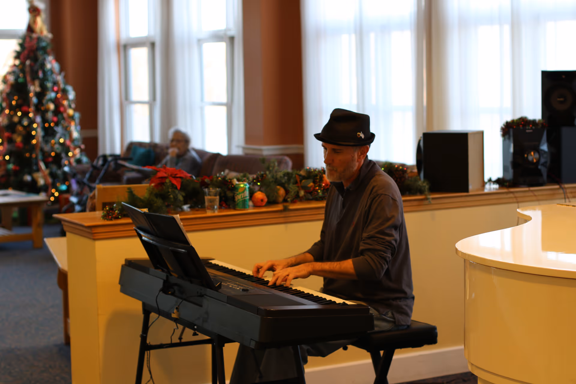 A man wearing a black hat plays an electronic keyboard in a decorated room with Christmas decorations, including a Christmas tree and garlands. In the background, an elderly person sits on a couch near large windows with white curtains.