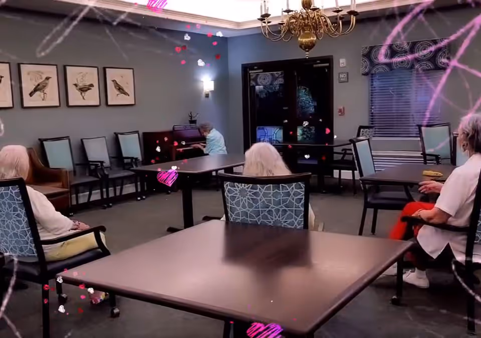 A common room in a senior living facility with several elderly individuals seated on chairs around tables. One person is playing a piano against the wall. The room has framed bird artwork on the wall, a chandelier, and a window with blinds and a valance.