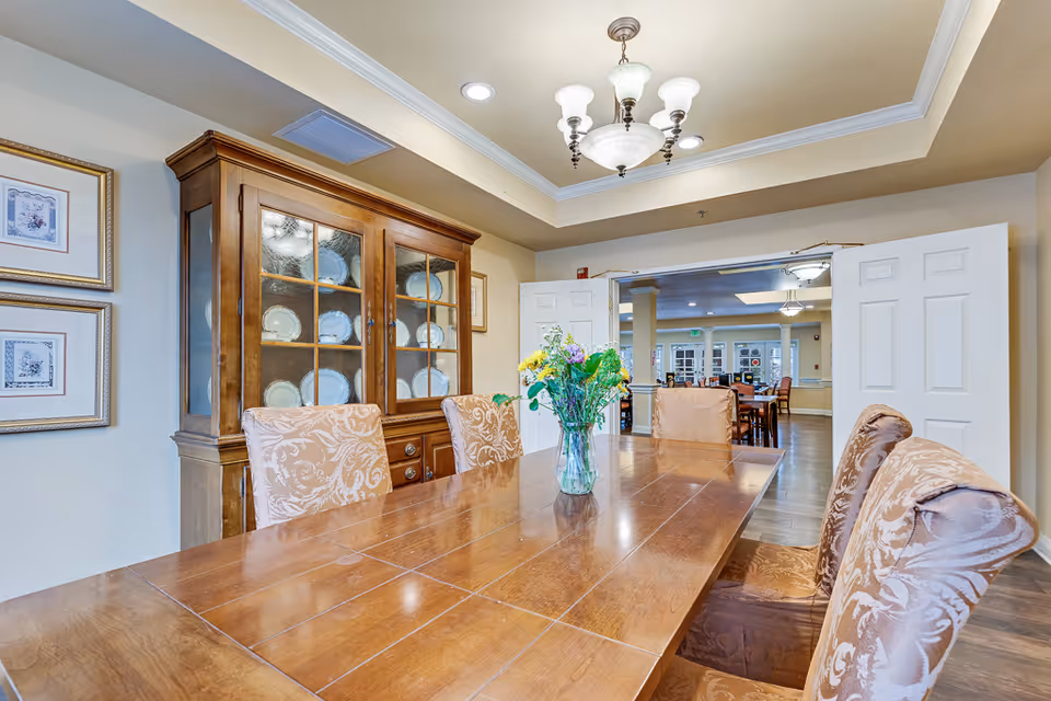A dining room with a wooden table and six upholstered chairs. A glass vase with colorful flowers is placed on the table. Behind the table is a wooden cabinet displaying plates. The room has beige walls, crown molding, and a chandelier hanging from the ceiling. Double doors open to another room with more tables and chairs.