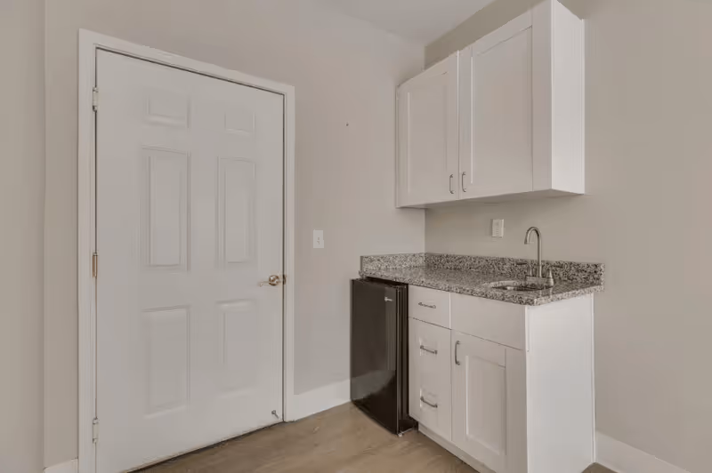 Small kitchenette area with white cabinets, a granite countertop, a stainless steel sink with a faucet, and a compact black refrigerator next to a closed white door in a room with light-colored walls and wood flooring.