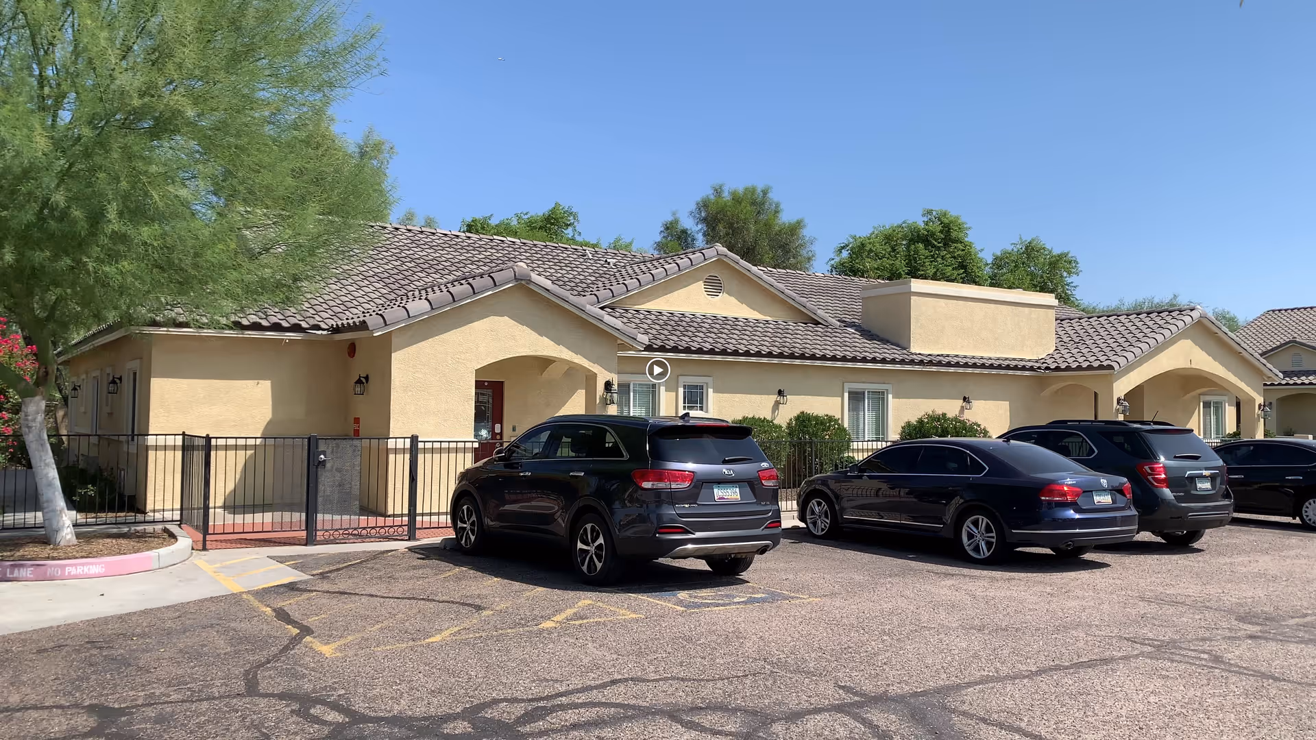 Exterior view of a single-story assisted living facility building with a beige stucco finish and tiled roof. Several cars are parked in front of the building in a paved parking lot. There are trees and shrubs around the building under a clear blue sky.