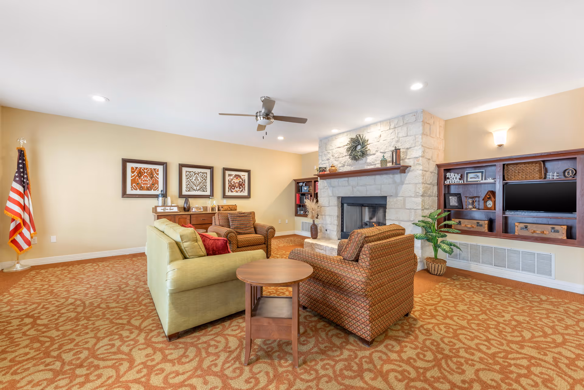 A cozy living room area with a green sofa, two patterned armchairs, a round wooden side table, and a stone fireplace with a wreath above it. The room has a patterned carpet, beige walls with three framed artworks, a ceiling fan, built-in wooden shelves with decorative items, and an American flag in the corner.