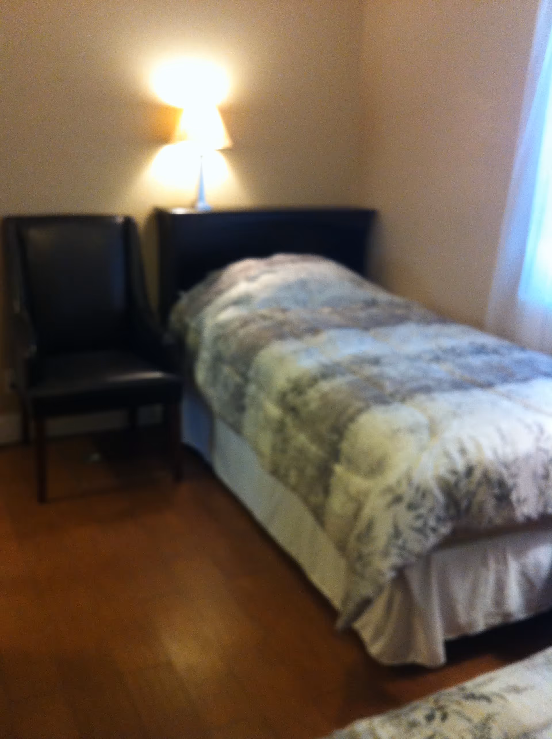 A small bedroom with a single bed covered with a patterned comforter. Next to the bed is a black chair and a nightstand with a lit lamp. The room has wooden flooring and a window with light curtains.