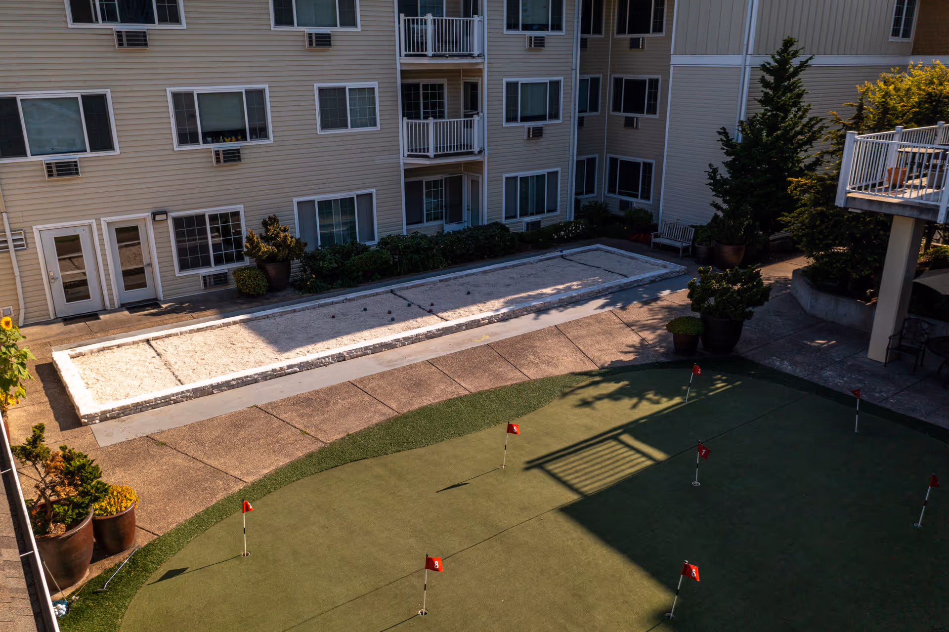 Outdoor courtyard area of a senior living facility with a putting green featuring several small red flags and a bocce ball court surrounded by potted plants and a multi-story building with balconies and windows.