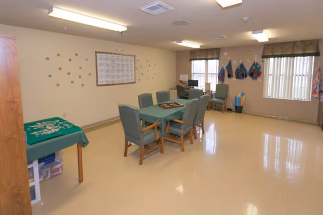 A spacious room with a table covered in a green tablecloth surrounded by six chairs. There is another small table with a green cover and some items on it to the left. The walls are beige with some decorations and a framed chart. Two windows with vertical blinds and valances are on the right side, and there are some hanging items and a container with pool noodles near the windows. The floor is a light beige color and the ceiling has fluorescent lights.