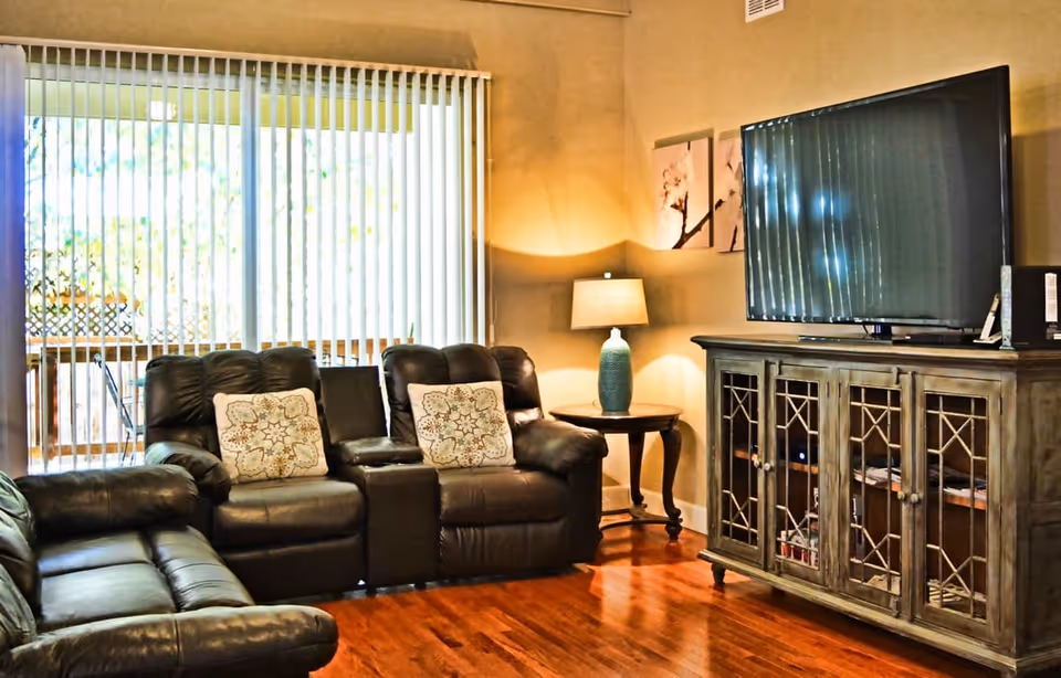 A cozy living room with dark leather recliner sofas, two decorative pillows, a wooden side table with a blue lamp, a large flat-screen TV on a wooden cabinet, and vertical blinds covering a sliding glass door that leads to an outdoor area.