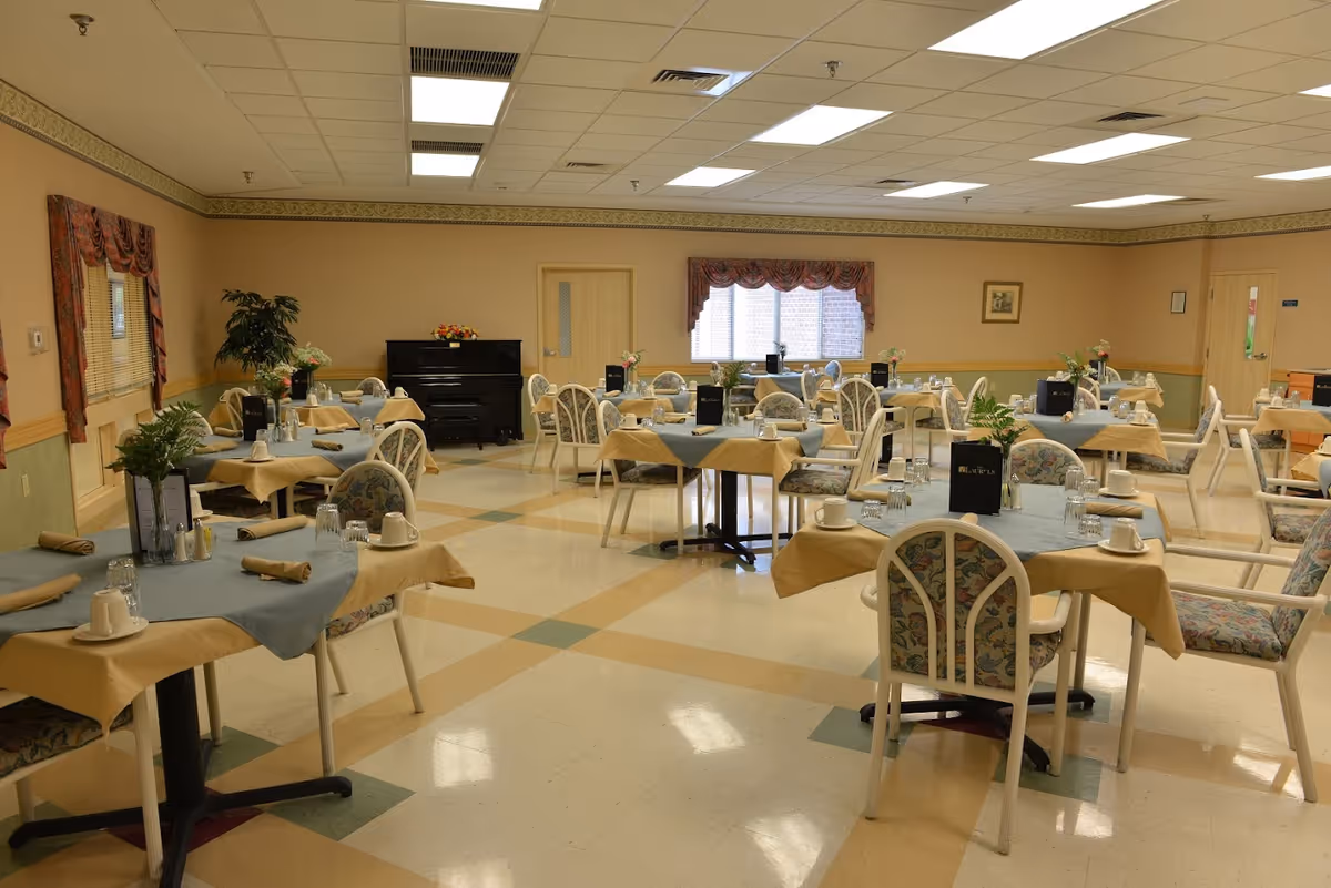 A spacious dining room with multiple tables covered in beige and blue tablecloths, each set with cups, glasses, napkins, and small flower arrangements. The room has floral-patterned chairs, beige walls with a green lower half, and windows with red curtains. A black piano is visible against the far wall.