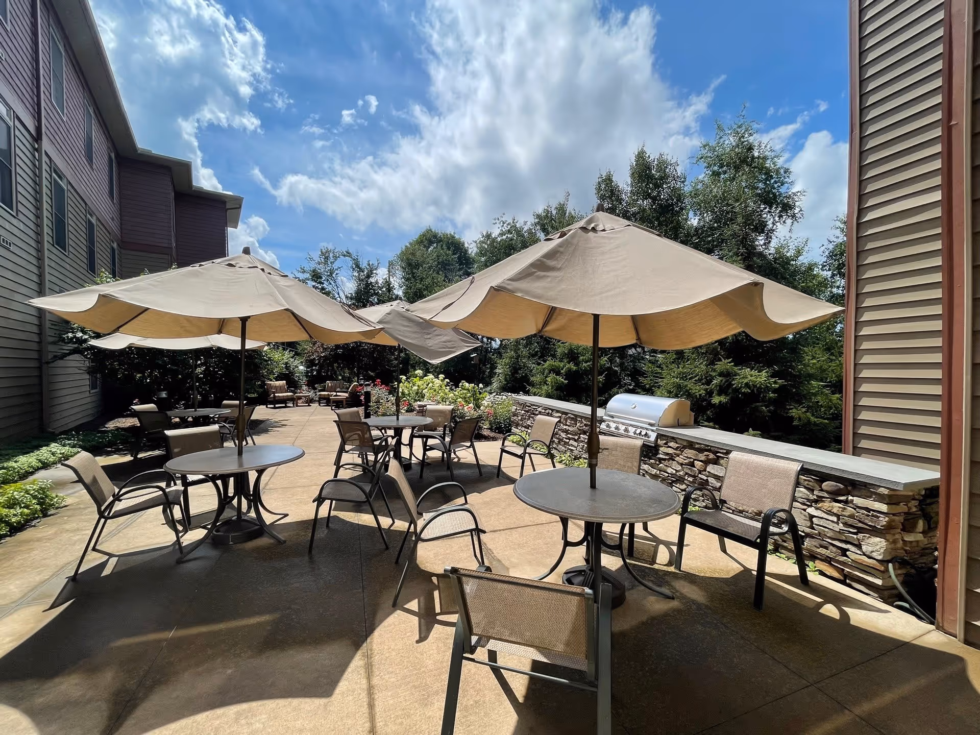 Outdoor patio area at The Pines Senior Living with round tables and chairs under beige umbrellas, a built-in stone grill, and greenery in the background under a partly cloudy blue sky.