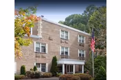 Front view of a three-story brick senior living building with a landscaped entrance and an American flag.