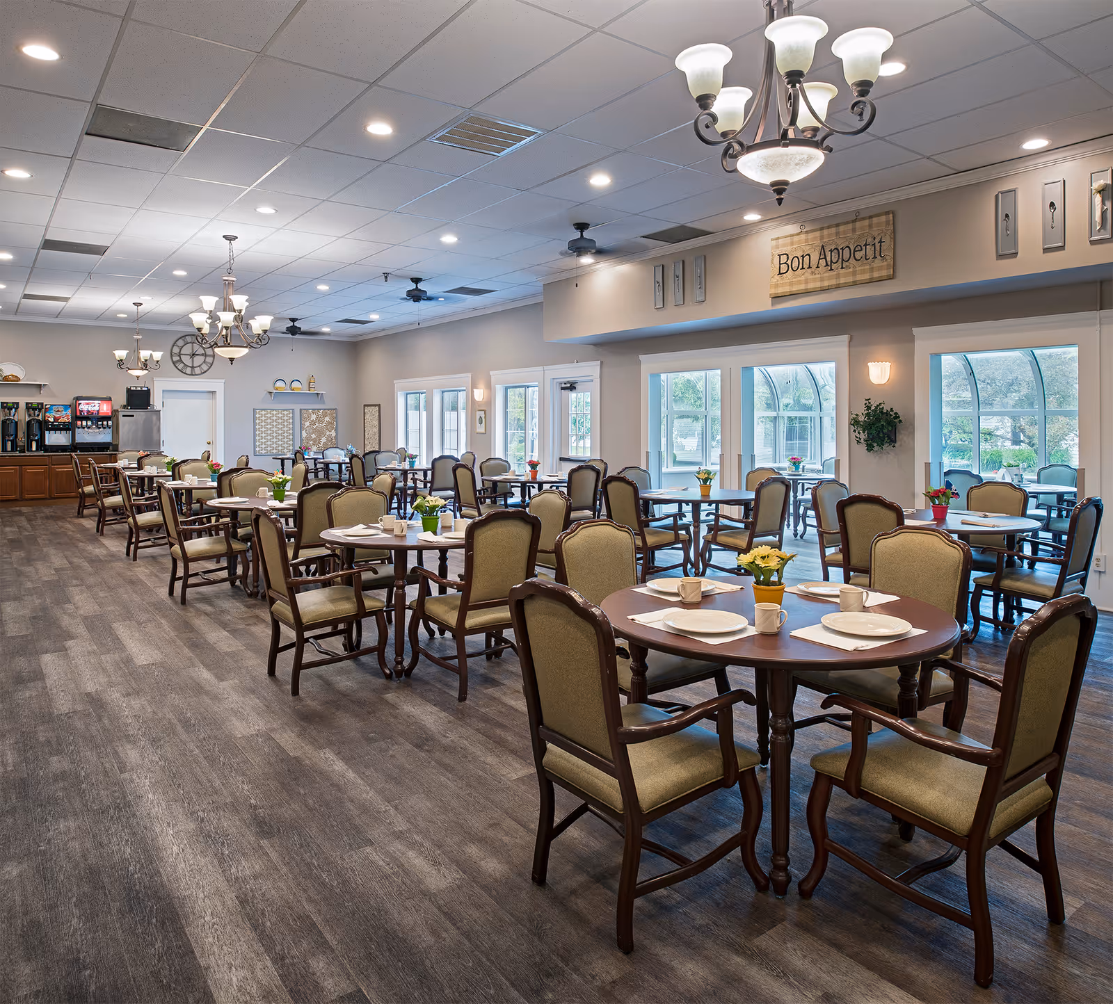 A spacious dining room with multiple round and rectangular tables set with plates, cups, and napkins. The room features wooden chairs with cushioned seats and backs, large windows letting in natural light, ceiling fans, and chandeliers. A sign on the wall reads 'Bon Appetit'. In the background, there is a beverage station with coffee machines and a soda dispenser.