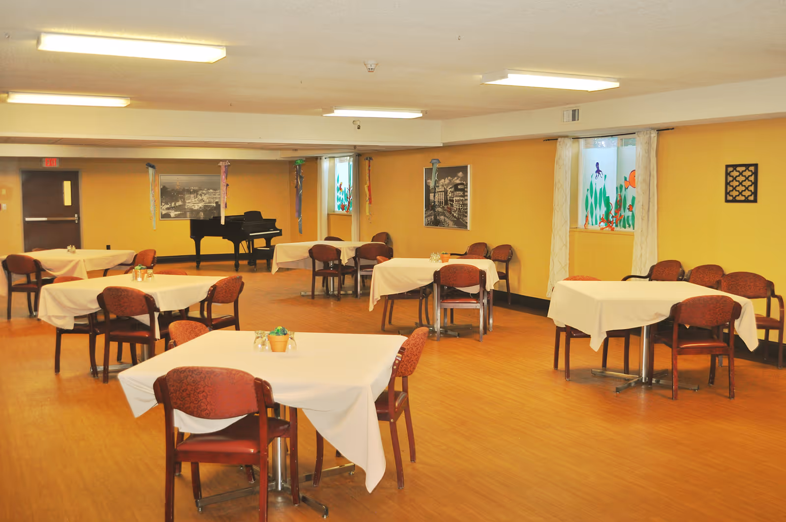 A spacious dining room with several square tables covered with white tablecloths, each surrounded by red cushioned chairs. The room has yellow walls, wooden flooring, and fluorescent ceiling lights. There are two windows with decorative curtains and colorful artwork on the glass. A black grand piano is positioned against the far wall, which also features framed black and white photographs and hanging decorations.