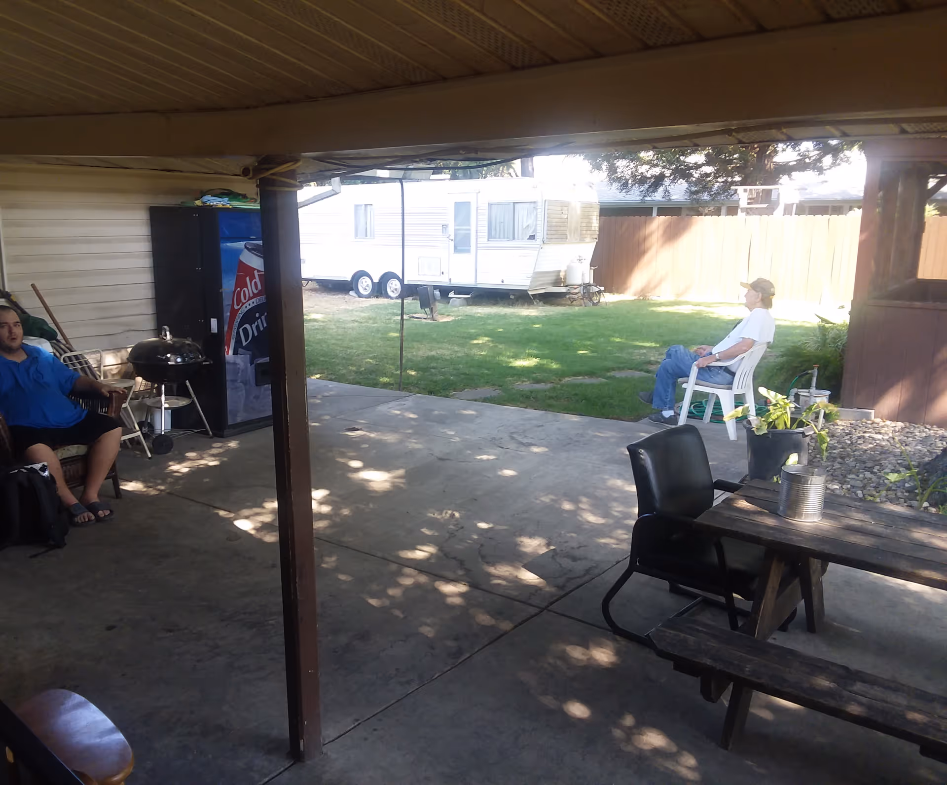 Covered patio area with concrete floor and wooden picnic table. Two men are seated, one on a plastic chair in the sun and the other on a chair in the shade. A vending machine and a charcoal grill are visible near the building wall. In the background, there is a grassy yard with a white trailer and a wooden fence.