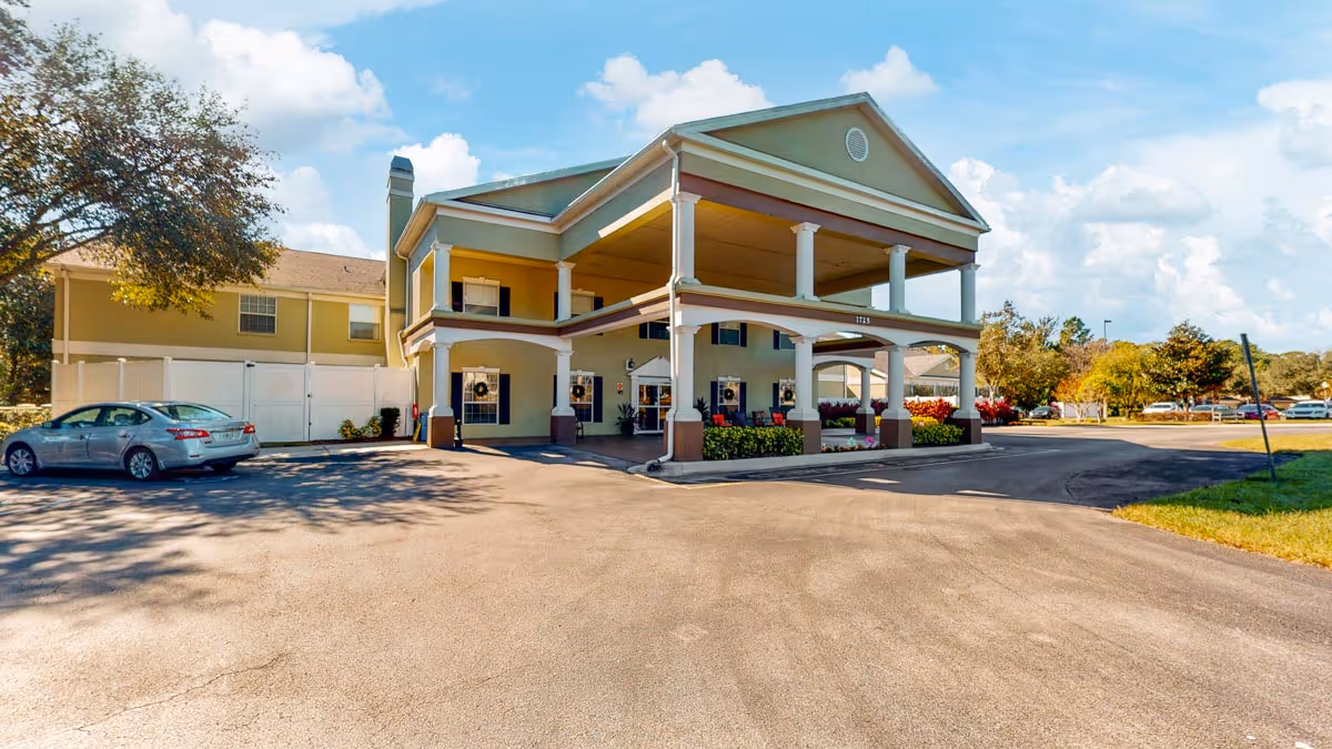 Front exterior of a two-story senior living facility with a covered porte-cochere, columns, and a parking area.