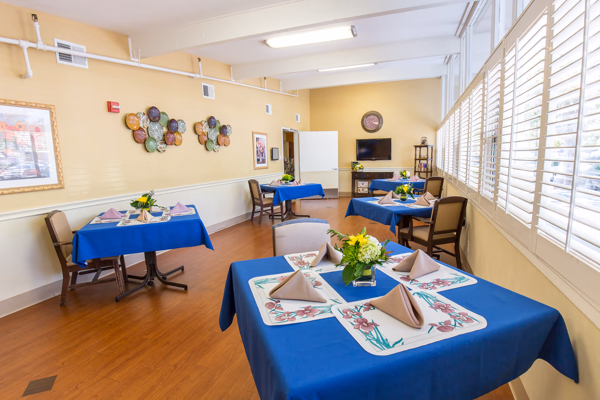A bright dining room with several tables covered in blue tablecloths, each set with folded beige napkins, floral placemats, and small flower arrangements. The room has wooden flooring, cream-colored walls with decorative plates and framed artwork, large windows with white shutters letting in natural light, and a TV mounted on the far wall.