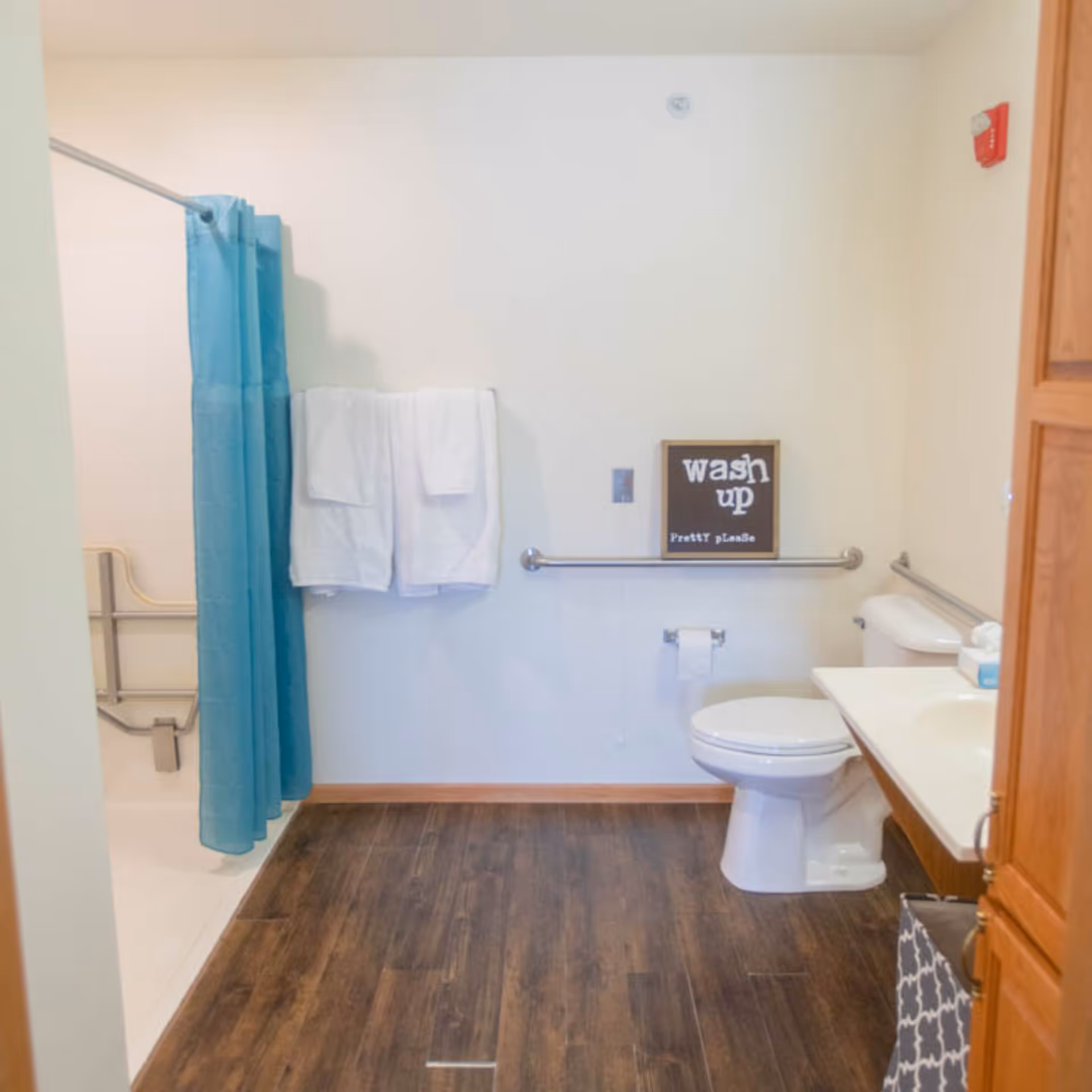 Accessible bathroom with a toilet and sink, grab bars, a shower with a blue curtain, towels on the wall, and a small "wash up" sign.