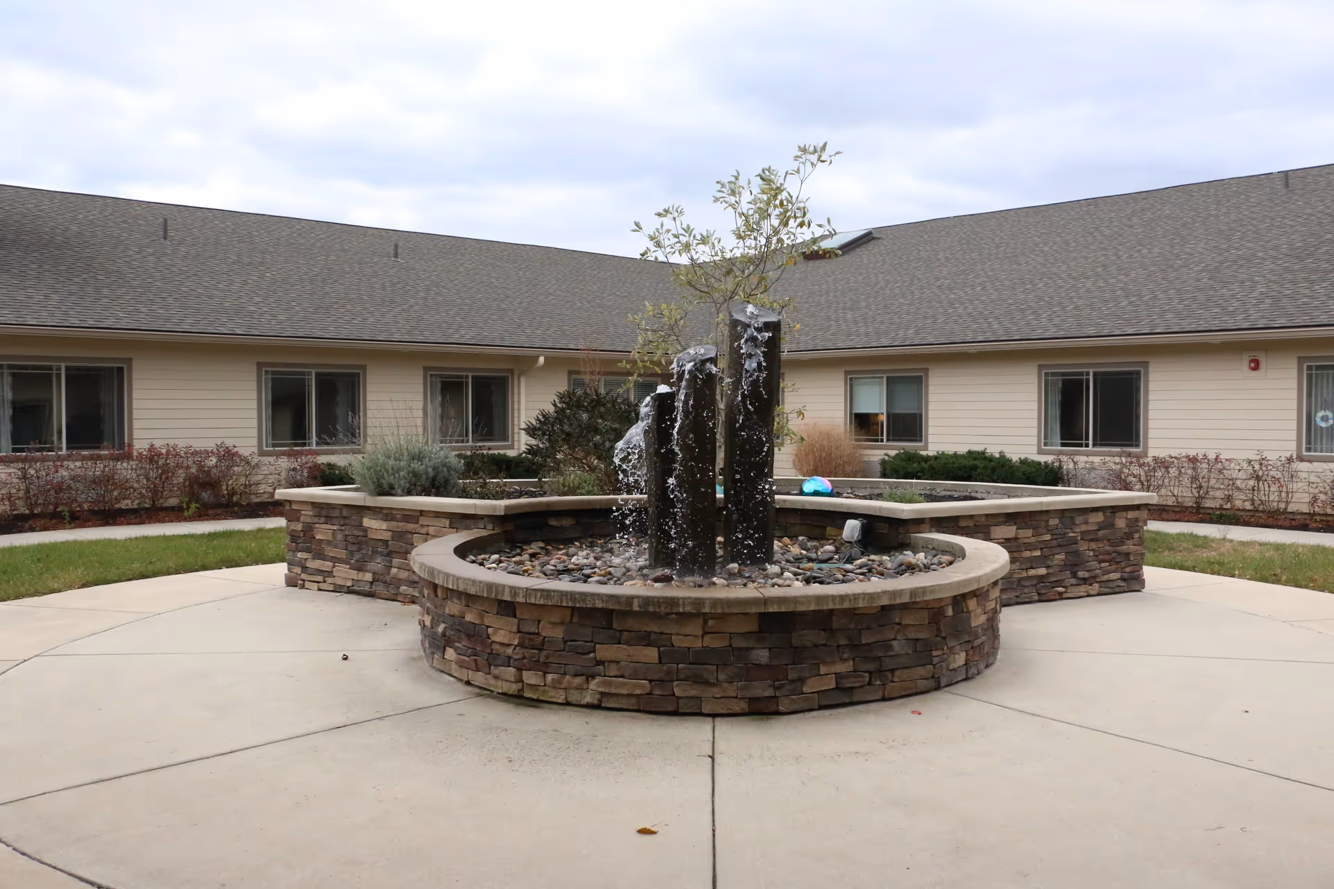 Outdoor courtyard area with a circular stone water fountain featuring three vertical stone pillars with water flowing over them. The fountain is surrounded by a paved walkway and landscaping with shrubs and small trees. The background shows a single-story building with beige siding and multiple windows under a cloudy sky.