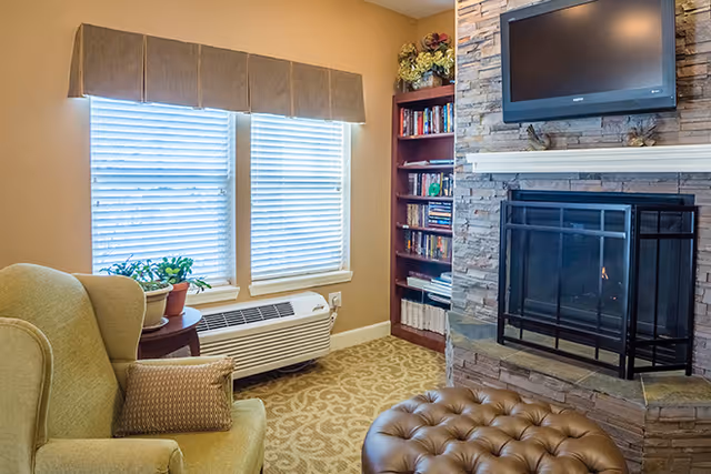 Cozy living room area with a green armchair and a brown pillow, a round ottoman, a stone fireplace with a black metal screen, a mounted flat-screen TV above the fireplace, a wooden bookshelf filled with books and decorative items, two windows with blinds and a beige valance, and a small table with potted plants next to the armchair.
