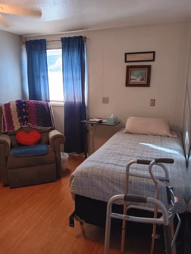 A small bedroom with a single bed covered in a plaid bedspread, a pillow, and a walker positioned at the foot of the bed. Next to the bed is a small table with a box of tissues and some electronic devices. There is a window with blue curtains letting in natural light. To the left of the window is a cushioned armchair with a colorful knitted blanket draped over the back and a red heart-shaped pillow on the seat. The floor is wooden, and two framed pictures hang on the wall above the bed.