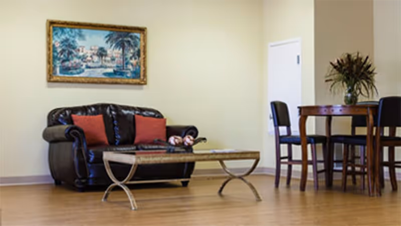 A cozy living room area with a dark leather loveseat adorned with two red cushions, a glass-top coffee table with decorative items, a wooden dining table with four chairs, and a vase with flowers on the table. A framed painting hangs on the light-colored wall.