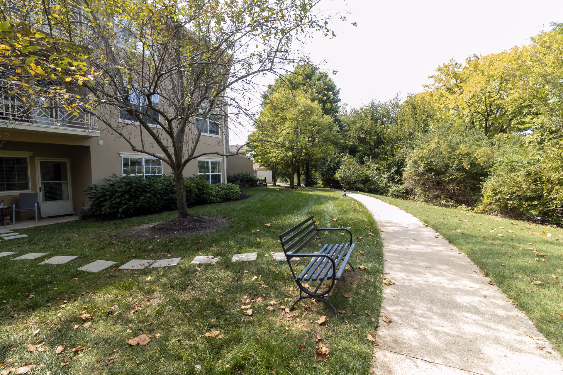 Outdoor area of a senior living facility with a concrete pathway curving through green grass and trees. A black metal bench is placed beside the pathway. The building with windows and a door is visible on the left side, along with some bushes and a tree with sparse leaves.