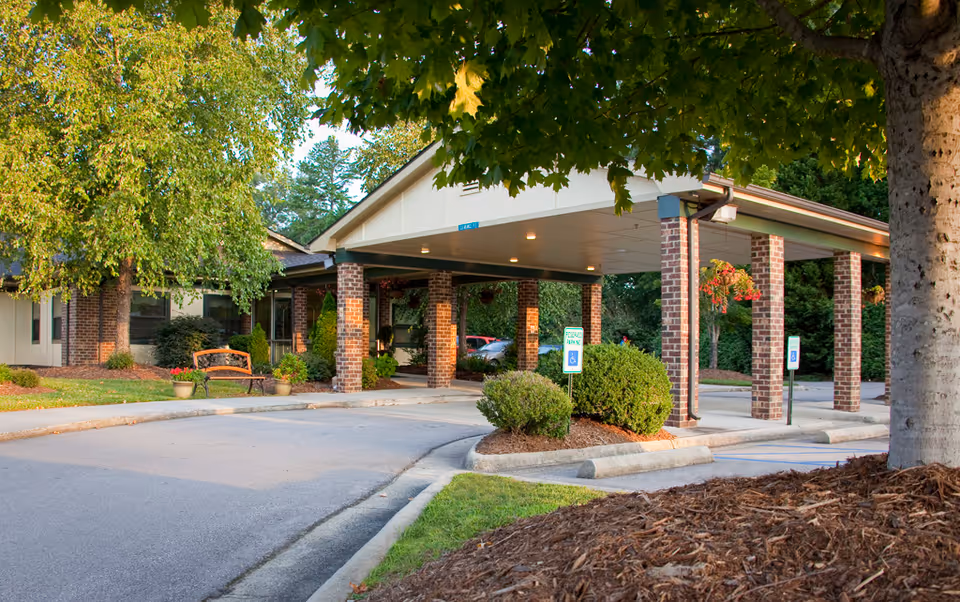 Covered brick-columned entrance and drive-up portico of a senior living facility with landscaping, a bench, and surrounding trees.