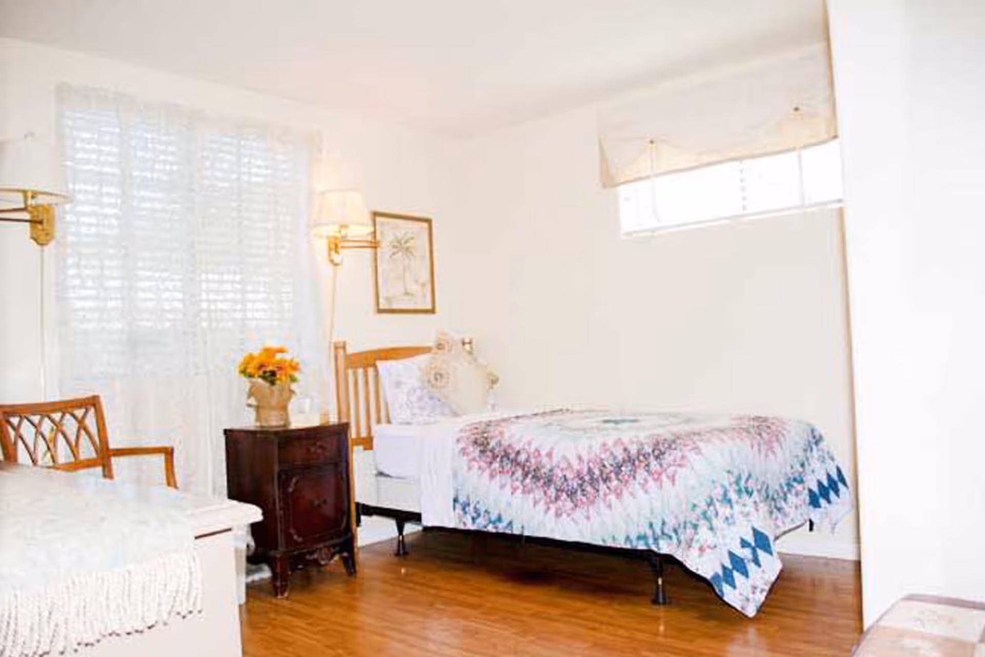 A bright bedroom with a single bed covered in a colorful quilt. Next to the bed is a dark wooden nightstand with a vase of yellow flowers. There is a wooden chair with a woven backrest near a window with white curtains. The room has light-colored walls, wooden flooring, and a framed picture on the wall above the nightstand.