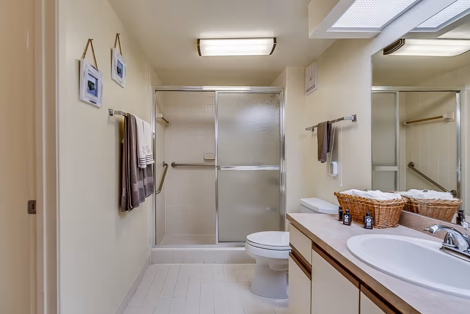 A clean bathroom with a glass-enclosed shower, a toilet, a sink with a countertop, and a large mirror. There are two framed pictures hanging on the wall, towels on towel racks, and a wicker basket with toiletries on the counter.