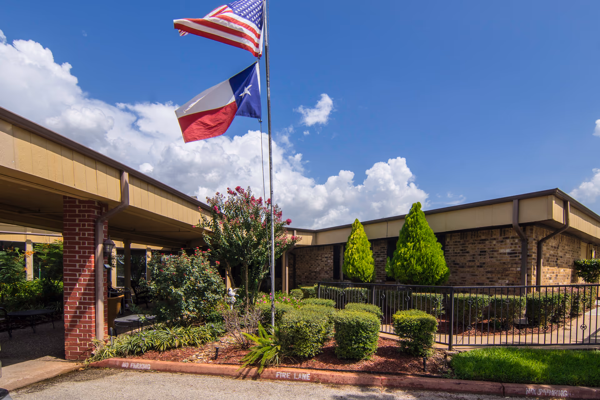 Front exterior of a single-story brick healthcare building with a covered entrance, landscaped shrubs, and American and Texas flags against a blue sky.