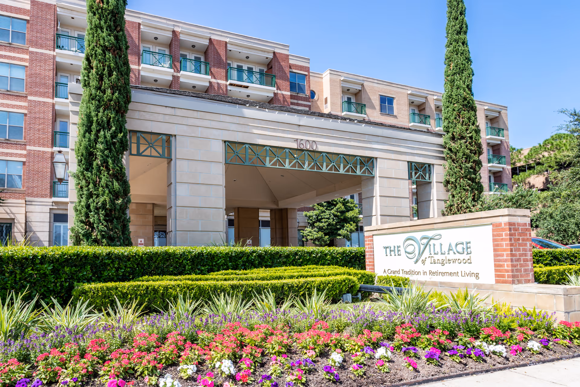 Exterior view of The Village of Tanglewood retirement living facility with a landscaped garden featuring colorful flowers and neatly trimmed bushes in front. The building has a brick and stone facade with balconies and tall trees flanking the entrance. A sign in front reads 'The Village of Tanglewood - A Grand Tradition in Retirement Living.'