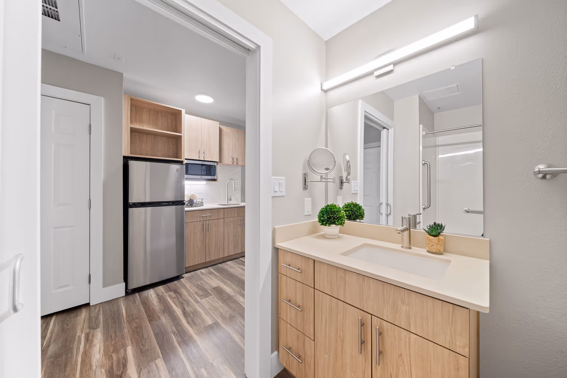 View of a modern bathroom vanity with a large mirror, light fixture, and small potted plants on the countertop. The bathroom opens into a kitchen area with wood cabinets, a stainless steel refrigerator, microwave, and sink. The flooring is wood-style laminate throughout.