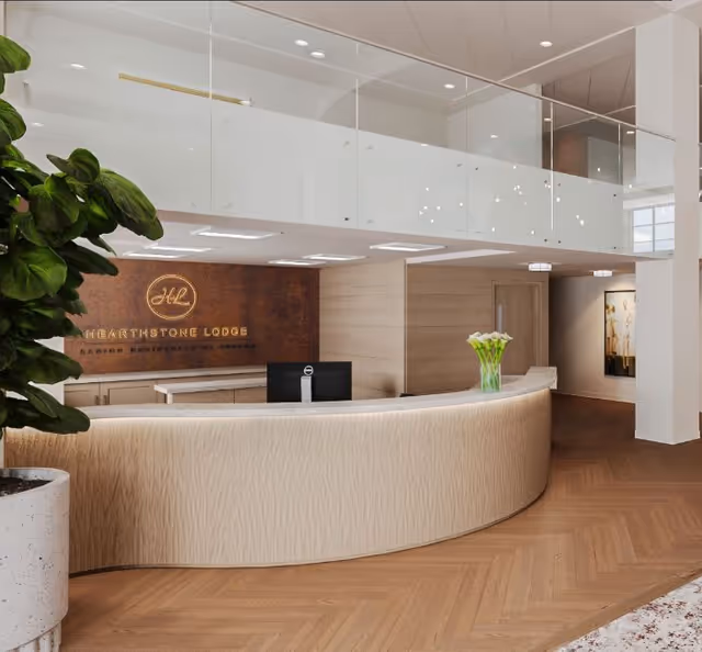 Modern senior living facility reception area with a curved beige front desk, a computer monitor, a vase with white flowers, wooden flooring, a large potted plant on the left, and a wall sign reading Hearthstone Lodge behind the desk.