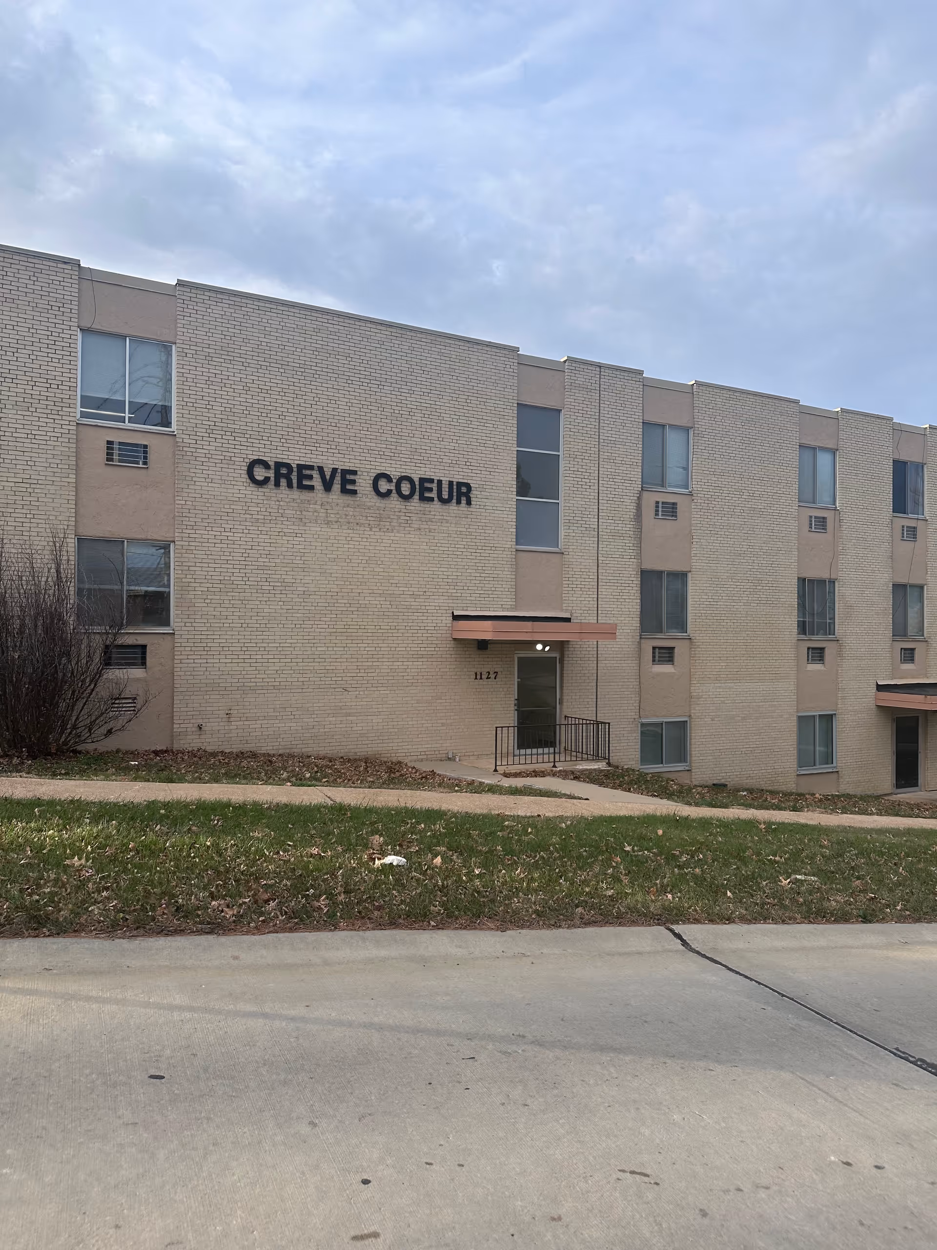 Exterior view of a beige brick building with the name 'CREVE COEUR' displayed on the wall. The building has multiple windows and two entrances with small awnings. There is a grassy area and a sidewalk in front of the building under a cloudy sky.