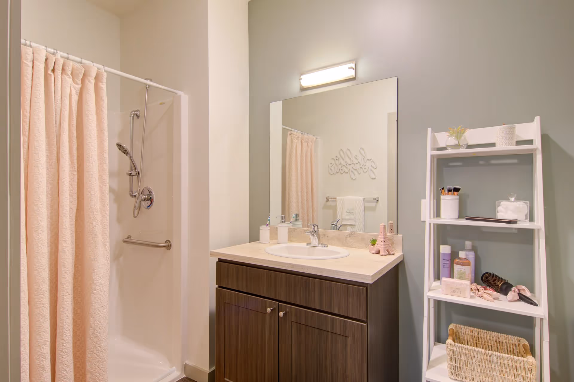 Small bathroom with a shower and pink curtain, a sink vanity with mirror, and a white shelving unit holding toiletries.