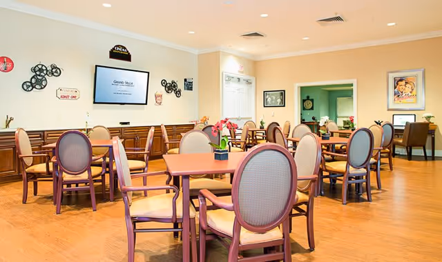 A bright and spacious dining room with multiple wooden tables and cushioned chairs arranged neatly. The walls are decorated with vintage movie-themed signs and framed pictures. A flat-screen TV is mounted on the wall displaying 'Grand Villa'. The room has wooden flooring and a computer workstation is visible in the background.