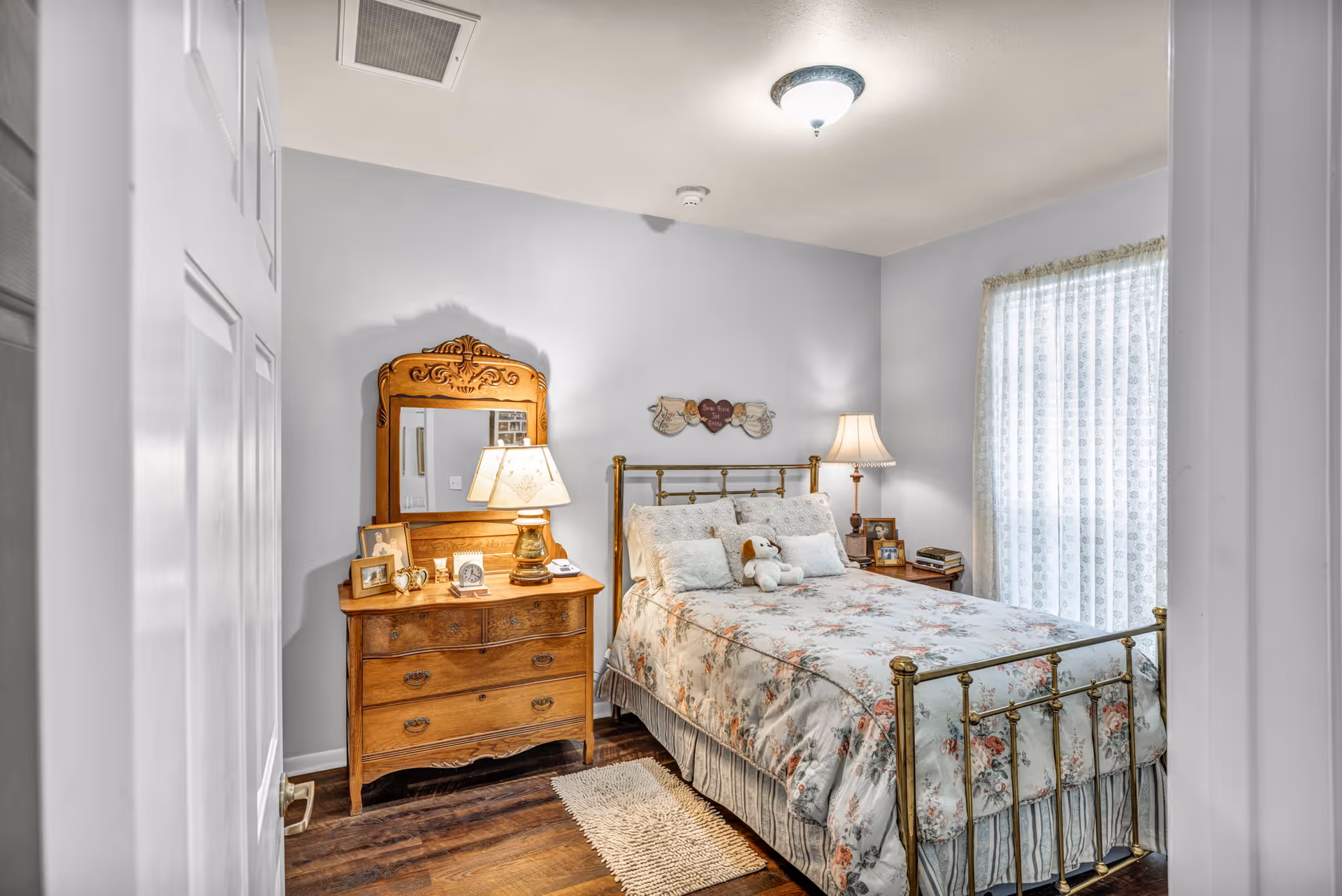 Cozy bedroom with a brass bed dressed in floral bedding, a wooden dresser with mirror and lamps, and a lace-curtained window.