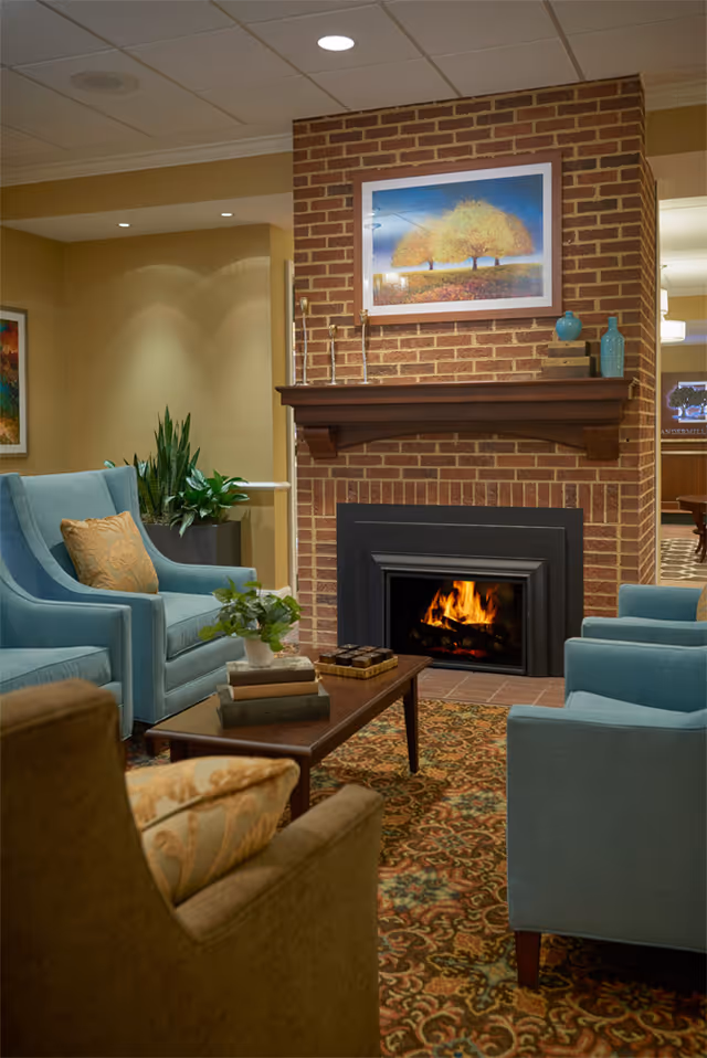 Cozy living room area with a lit fireplace set in a brick wall. The room features blue upholstered armchairs with decorative pillows, a wooden coffee table with books and a plant, a patterned carpet, and framed artwork above the fireplace. There are also some decorative vases on the mantel and plants in the background.