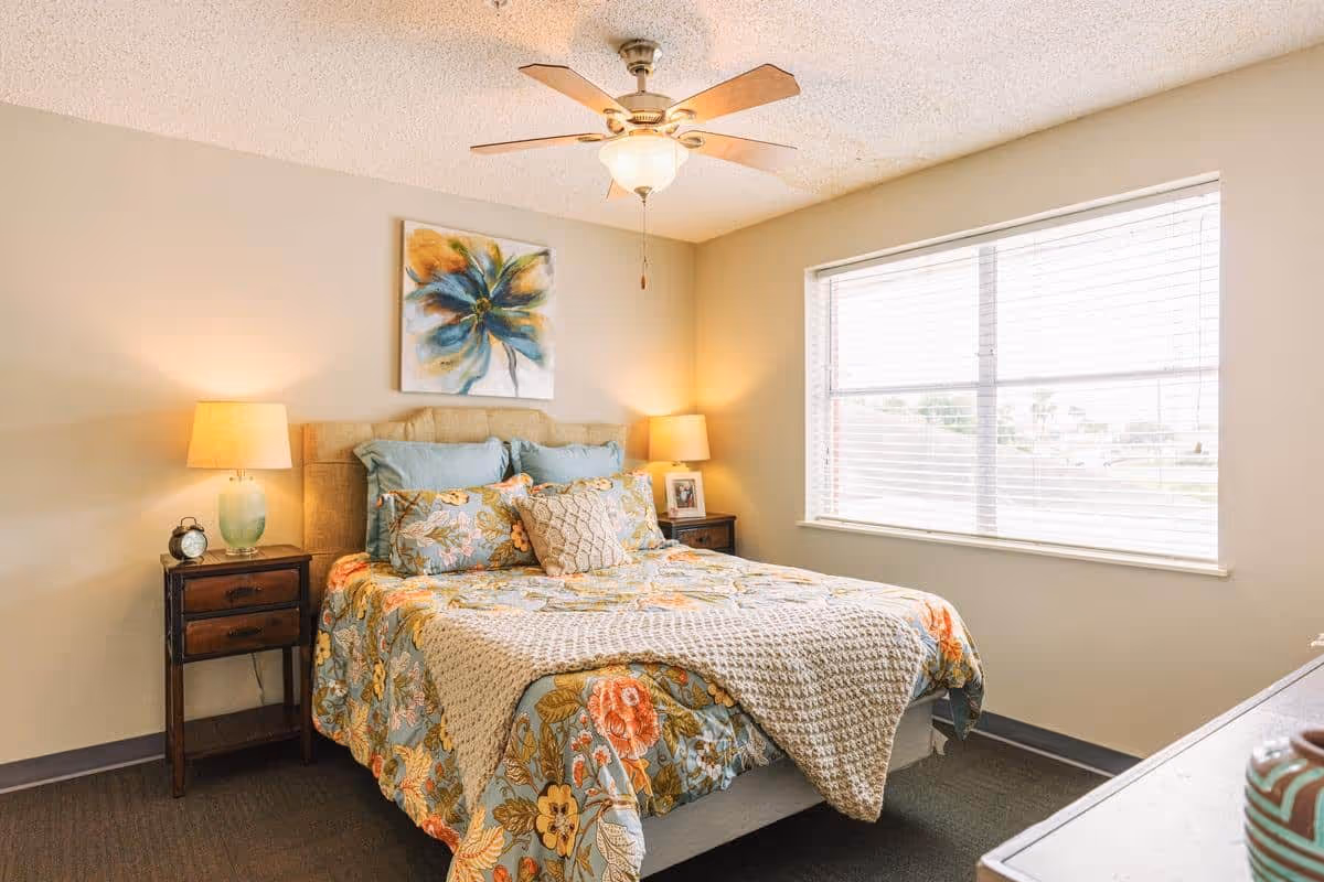 A cozy bedroom with a bed covered in a floral patterned comforter and multiple pillows. There are two wooden nightstands on either side of the bed, each with a lamp. A ceiling fan with a light is mounted on the ceiling. A large window with blinds allows natural light into the room. A colorful abstract flower painting hangs above the bed.
