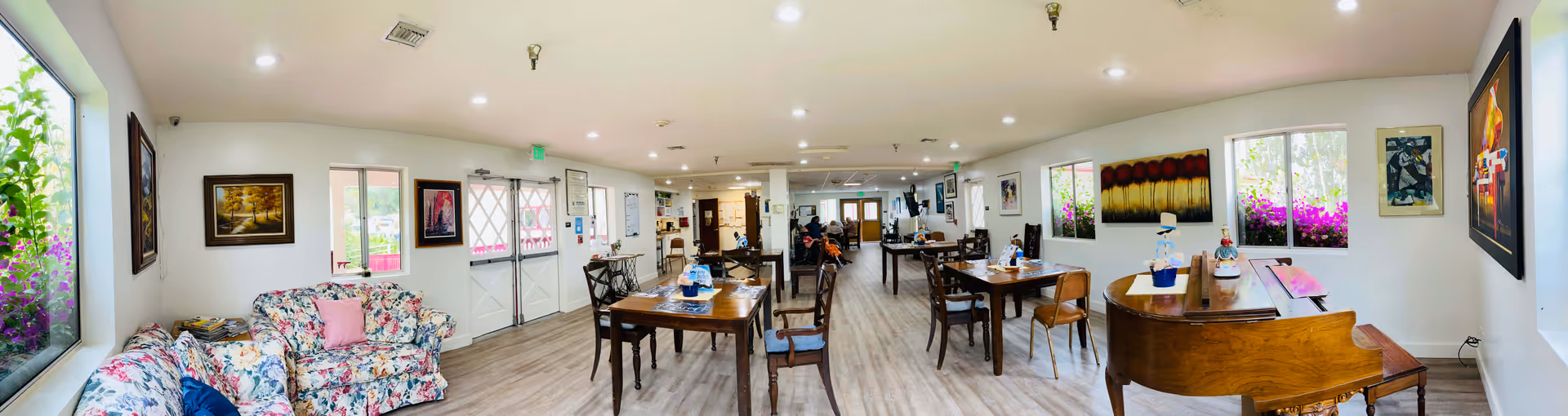 A spacious, well-lit common area in a senior living facility featuring floral patterned sofas on the left, multiple wooden tables and chairs arranged throughout the room, and a grand piano on the right. The walls are decorated with various paintings and large windows allow natural light to brighten the space.