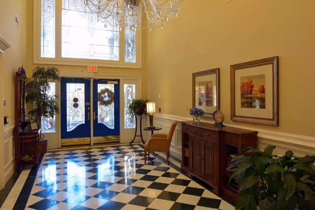 Bright entry foyer with blue double doors, a chandelier, black-and-white checkered floor, and a wooden sideboard with framed art.
