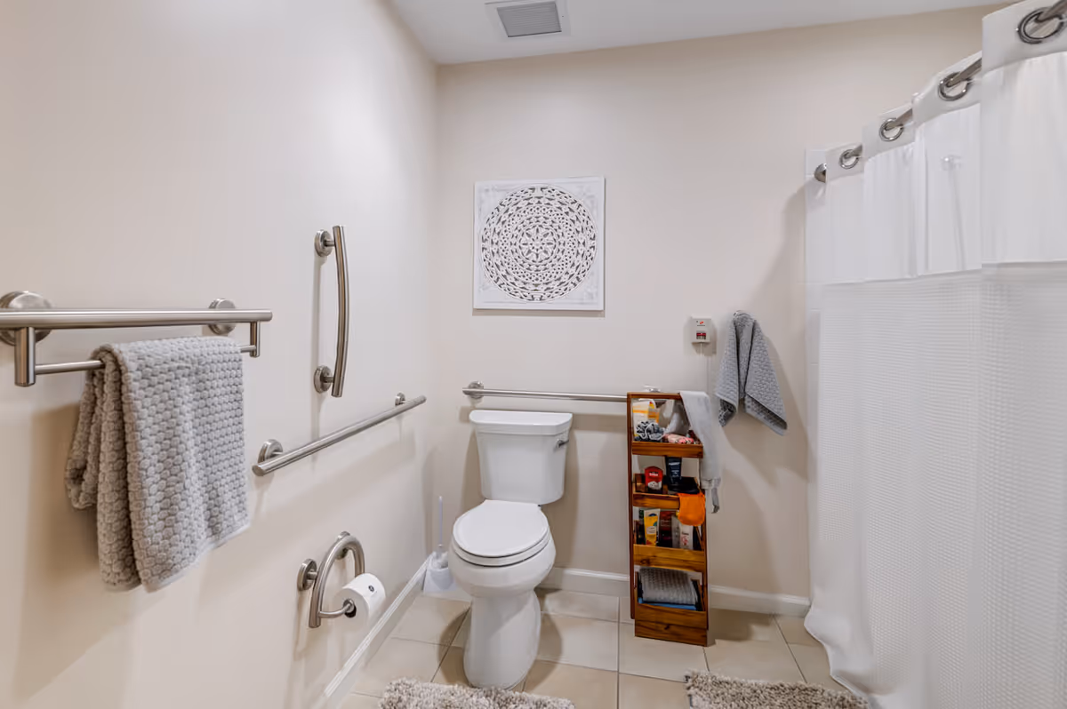 A clean and accessible bathroom with beige walls and tiled floor. It features a white toilet with grab bars on the walls around it for support. There is a towel rack with a gray textured towel, a small wooden shelf holding toiletries and towels, a white shower curtain, and a decorative white wall art piece above the toilet.