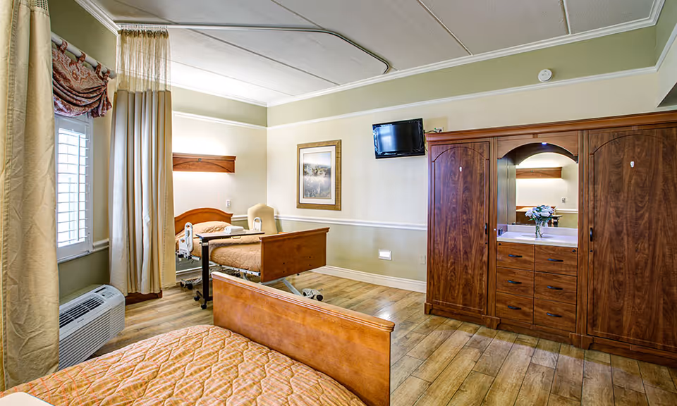 Bright furnished senior bedroom with two wooden beds, a large wardrobe with mirror, a wall-mounted TV, and windows with curtains.
