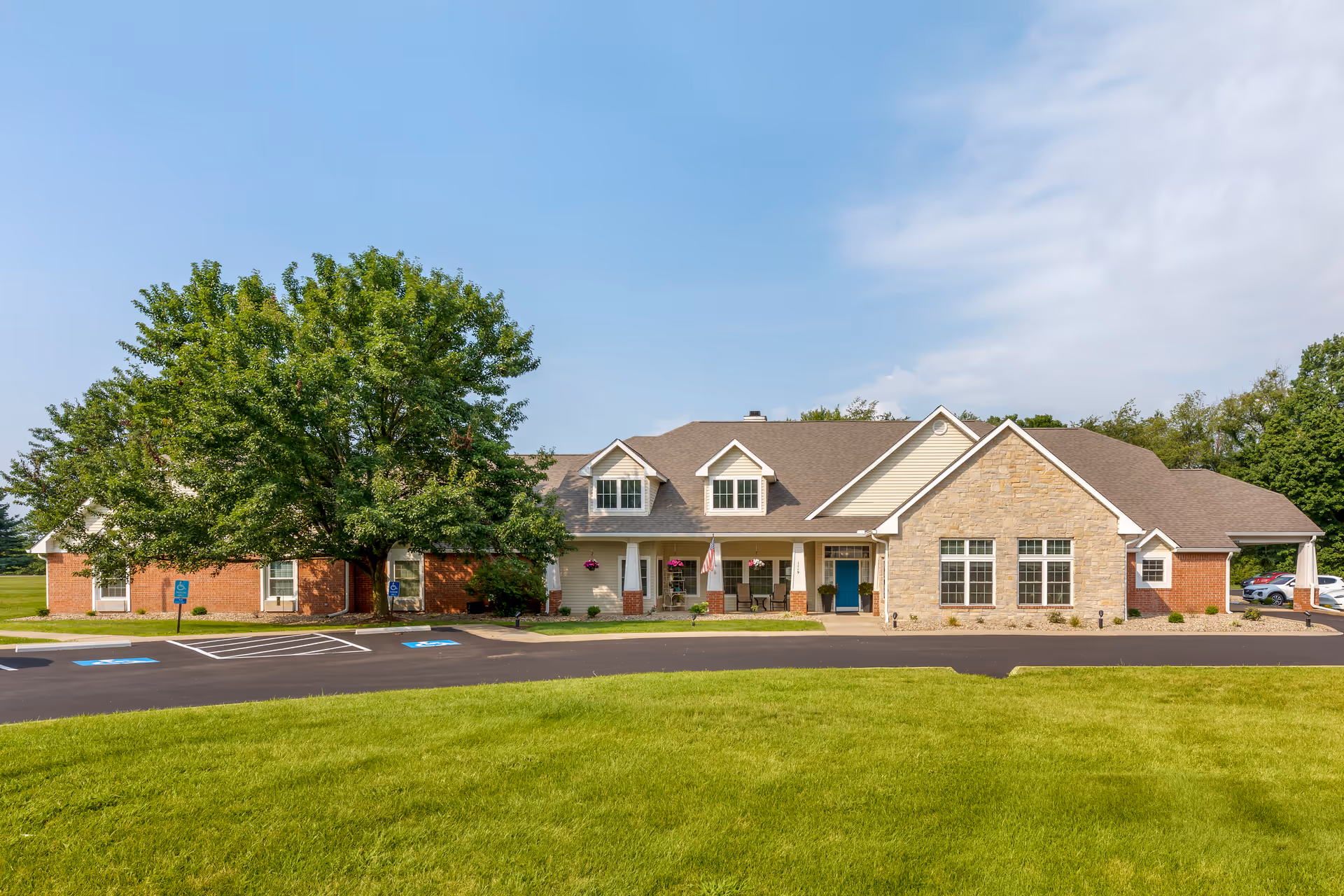 Front exterior view of a single-story senior living facility building with a combination of brick, stone, and siding. The building has a covered entrance with a blue door, several windows, and dormer windows on the roof. There are hanging flower baskets and an American flag near the entrance. The foreground features a well-maintained lawn and a paved parking area with handicap parking spaces. Trees and a clear blue sky are visible in the background.
